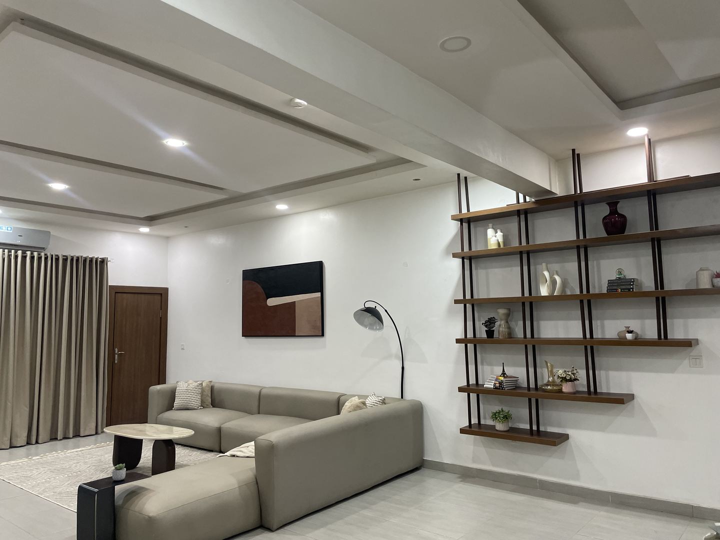 Wide view of a neutral living room with sectional sofa, sculptural coffee tables and open shelving